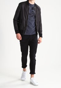 Svart bomberjacka med ribbat krage över en marinblå t-shirt med grafik. Svart slim fit-jeans och vita sneakers fulländar looken.