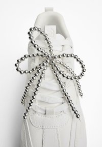 Vita sneaker med texturerad läderöverdel, med en silverfärgad pärlrosett som accent vid snörningen och platta bomullsnöre.