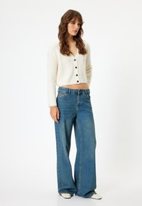 Cremefarbener Rippstrick-Cardigan mit V-Ausschnitt und Knopfverschluss, kombiniert mit hochgeschnittenen, weiten blauen Jeans aus Denim. Sauberer, lässiger Stil mit heller Waschung.