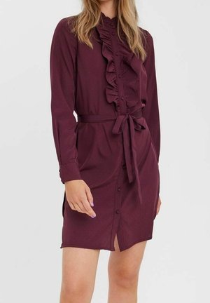 Vrouw draagt een bordeauxrode jurk met lange mouwen, knopen aan de voorkant, ruches en een ceintuur in de taille.