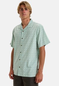Camicia a maniche corte di colore verde menta con motivi floreali bianchi, vestibilità rilassata e colletto. Bottoni marroni decorano la parte anteriore.