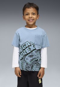 Šypsosi jaunas berniukas, vilkintis sluoksniuotas mėlynai ir baltai ilgas rankoves marškiniais su dinozaurų piešiniu ir juodomis šortais, prieš pilką foną.