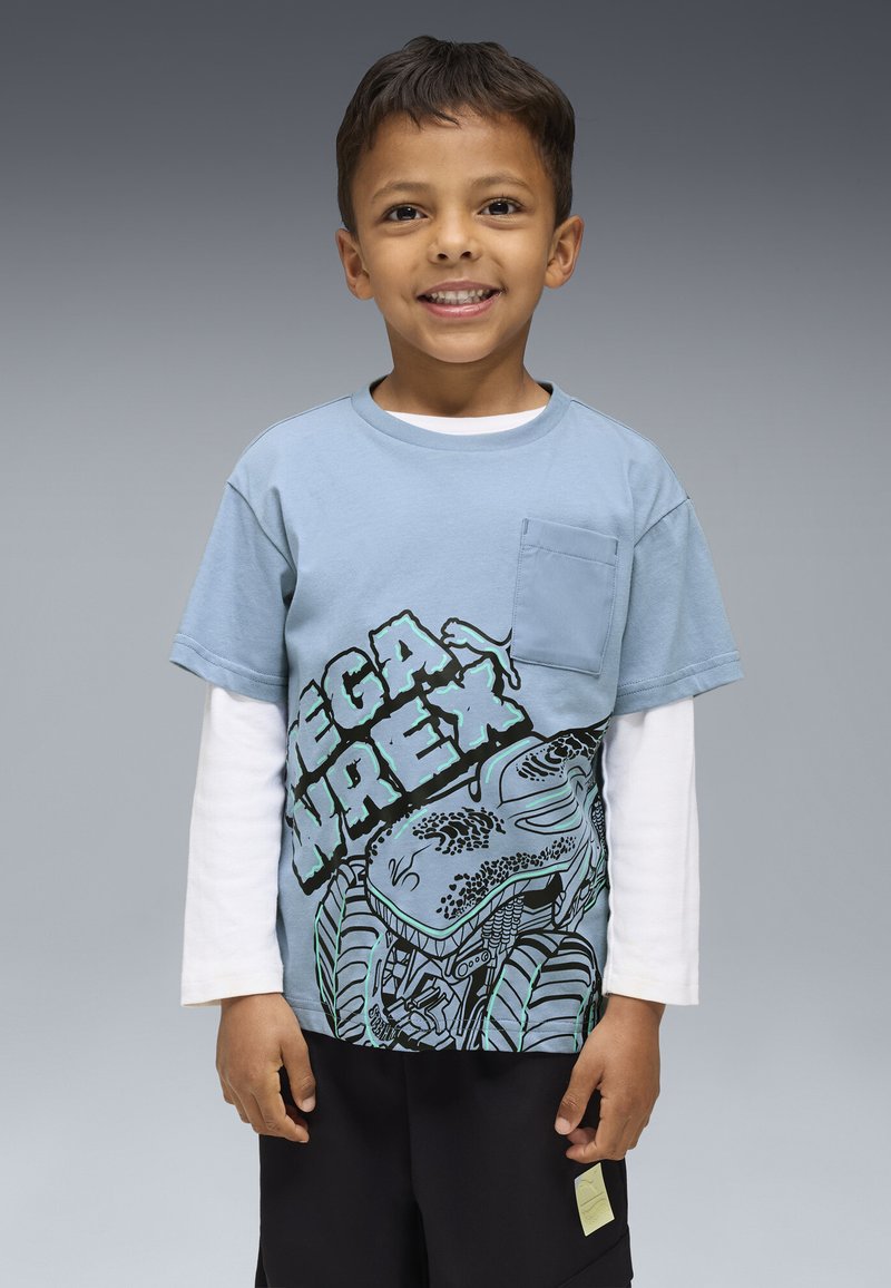 Šypsosi jaunas berniukas, vilkintis sluoksniuotas mėlynai ir baltai ilgas rankoves marškiniais su dinozaurų piešiniu ir juodomis šortais, prieš pilką foną.