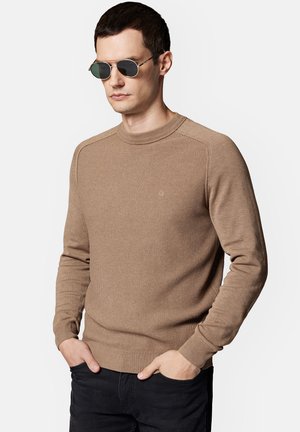 Brązowy sweter z prążkowanej tkaniny, z okrągłym dekoltem, dopasowanymi rękawami i subtelnym detalem logo. Stylizowany z ciemnymi dżinsami i okularami przeciwsłonecznymi.