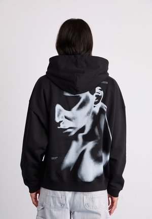 Black hoodie with a large gray graphic of a face on the back. Features a hood and oversized silhouette. Material appears soft with a smooth finish.