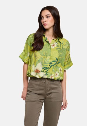 Mujer con camisa de manga corta verde con botones, estampado grande de flores y hojas blancas y amarillas, combinada con pantalones caqui de cintura alta.
