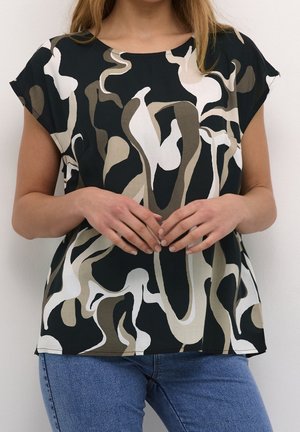 Vrouw draagt een blouse met korte mouwen met een abstract patroon in zwart, wit en beige, gecombineerd met een blauwe spijkerbroek, handen gevouwen op de taille.