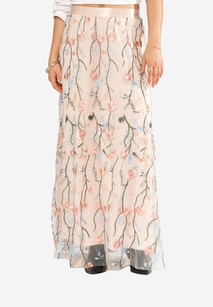 Jupe maxi beige avec broderie florale rose, bleue et verte, portée avec des chaussures noires et un haut court blanc.