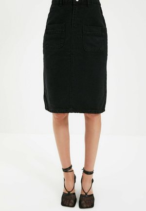 Zwarte knielange rok met opgestikte zakken aan de voorkant, gedragen met zwarte mesh hoge hakken met bandjes, op een witte achtergrond.