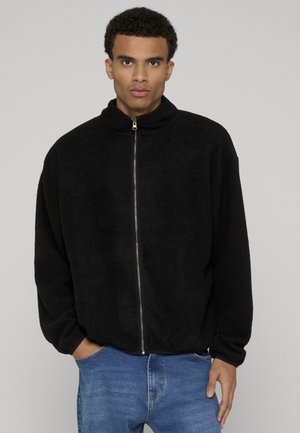 Młody mężczyzna z krótkimi kręconymi włosami, ubrany w czarną polarową kurtkę na zamek i niebieskie dżinsy, stojący z jedną ręką w kieszeni na jednolitym tle.