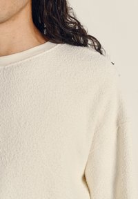 Vit texturerad sweatshirt med rund halsringning och något sänkta axlar. Materialet verkar mjukt och mysigt, med minimala sömmar synliga.