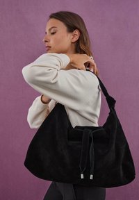Sort ruskinds hobo-taske med snørelukning, blød tekstur, rund form og tofarvede remme, holdt mod en lilla baggrund.