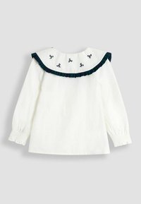 Weiße Bluse mit langen Ärmeln, rundem Ausschnitt, grün-blau gestreiftem Volant und gestickten schwarzen Schleifen an der Passe. Glatte Textur.