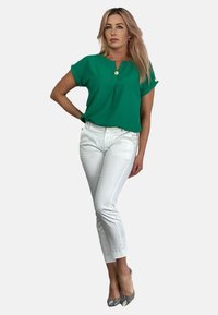 Zielona bluzka z krótkim rękawem i kołnierzykiem, z złotym guzikiem, połączona z białymi dopasowanymi spodniami i wzorzystymi szpilkami.