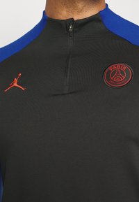 Nahaufnahme eines schwarzen Sporttrikots mit blauen Schultern, halb-zip Kragen, roten Paris Saint-Germain und Jordan-Logos auf der Brust.