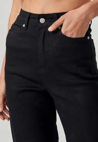 Visoko pasne črne kavbojke iz denima z gladko teksturo, kontrastnim šivom in sprednjimi žepi. Klasična kovinska zaponka je vidna.