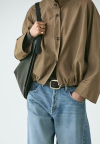 Person trägt eine braune, geknöpfte Jacke, blaue Jeans, schwarzen Gürtel mit silberner Schnalle, trägt schwarze Umhängetasche, eine Hand in der Tasche, Ringe an den Fingern.