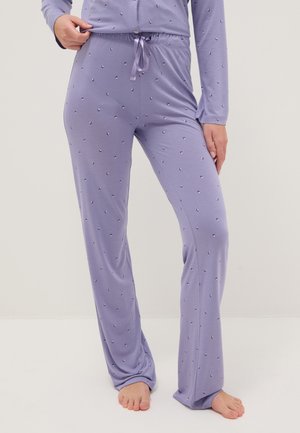 Persona vistiendo pantalones de pijama color lila claro con un pequeño patrón disperso, de pie descalza sobre un fondo liso.