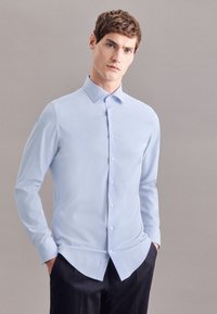 Camicia azzurro chiaro a maniche lunghe con colletto a punta, chiusura frontale con bottoni, tessuto liscio e orlo leggermente curvo, abbinata a pantaloni scuri.