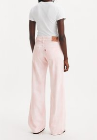 Une personne portant un jean large rose clair et une chemise blanche ajustée à manches courtes, vue de dos sur fond blanc.