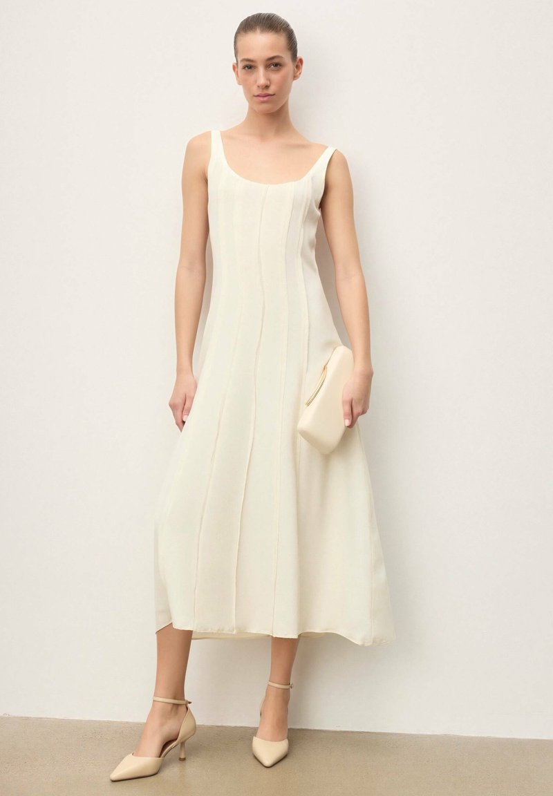 Femme portant une robe midi crème sans manches avec des coutures, tenant une pochette assortie, et chaussée de talons beige à bride à la cheville, posant devant un mur blanc.