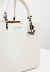 Vit läderhandväska med en textured finish, utrustad med korta handtag, en kedjeaccent och en silverfärgad logocharm. Diskret branding på sidan.