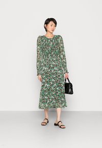 Vestido floral de mangas longas com padrão verde, branco e preto. Tecido transparente, cintura ajustada e saia fluida. Bolsa preta e sandálias rasas.