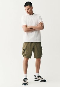 T-shirt blanc à manches courtes, shorts cargo vert olive avec poches, chaussettes blanches et baskets noires/grises. Design simple, style décontracté.
