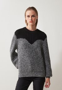 Strikket genser med en svart ribbestrikket bærestykke og en teksturert grå kropp. Designet inkluderer et chevron-mønster ved halsen.