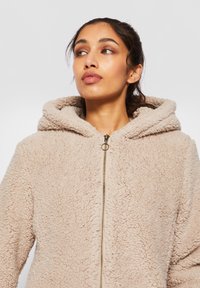 Kvinne med mørkt hår bundet bak, iført en beige, fluffy hettegenser med glidelås, ser litt opp og til siden.