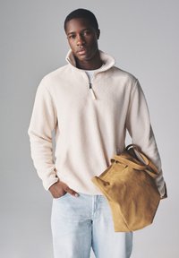 Jasnobeżowy polarowy pulower z zamkiem przy dekolcie, zestawiony z jasnymi jeansami. Osoba trzyma brązową torbę typu tote ze skóry zamszowej.