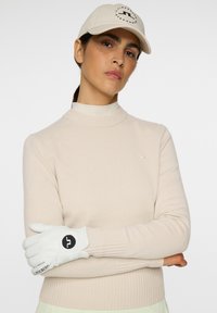 Maglione beige a costine, guanti bianchi con logo nero e cappellino chiaro con accentuazione grafica. Texture liscia, design aderente e colori neutri.