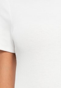 Gros plan d'une personne portant un t-shirt blanc à manches courtes côtelé, montrant l'épaule et la partie supérieure du torse.