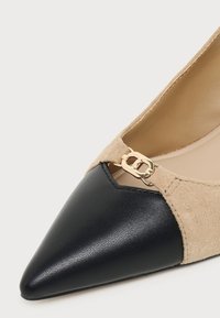 Chaussure plate à bout pointu avec devant en cuir noir, côtés en daim beige et petite boucle décorative dorée près du bord supérieur.