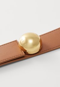 Ruskea nahkahihna, jossa on suuri, heijastava kultainen pyöreä metalliosuus, johon on kaiverrettu sana "FURLA".