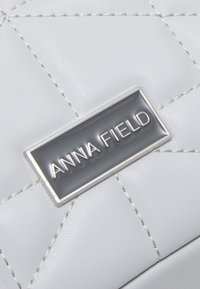 Hvid quiltet syntetisk lædertaske med sølvtråd og en rektangulær grå metalplade med teksten "ANNA FIELD."