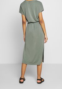 Mujer vestida con un vestido midi verde de mangas cortas y una abertura lateral, usando sandalias negras, de pie de espaldas a la cámara sobre un fondo liso.