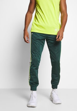 Mand iført mørkegrønne træningsbukser med hvide side logoer, gul stribe, neongult ærmeløst shirt og hvide atletik sko, der står.