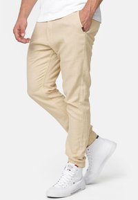 Pantalones beige ajustados con puños elásticos, tejido texturizado, bolsillos laterales y una pequeña etiqueta marrón en la cinturilla, combinados con zapatillas blancas de caña alta.