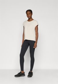 Μπεζ ανοιχτόχρωμο τοπ με κοντά μανίκια συνδυασμένο με μαύρα κολάν. Το σύνολο έχει χαλαρή εφαρμογή και λεία, ελαστική υφή. Μαύρα αθλητικά παπούτσια ολοκληρώνουν την εμφάνιση.