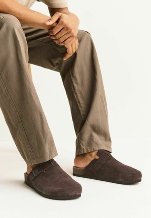 Person sitzt mit verschränkten Händen, trägt braune Slip-on Wildlederschuhe und beige Hosen vor einem einfarbigen weißen Hintergrund.