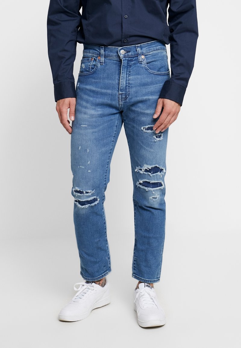 Ljusblå slitna jeans med revor och en tapered passform, kombinerat med vita sneakers och en mörk knapp-up skjorta.