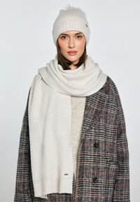 Kremowa dzianinowa czapka z pomponem, do kompletu miękki szal ułożony na teksturowanym szaro-brązowym płaszczu w kratę z czarnymi guzikami.