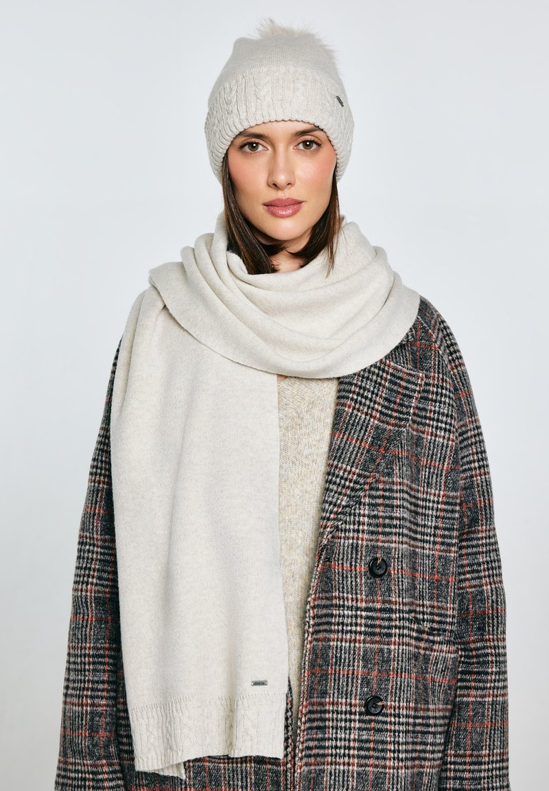 Kremowa dzianinowa czapka z pomponem, do kompletu miękki szal ułożony na teksturowanym szaro-brązowym płaszczu w kratę z czarnymi guzikami.