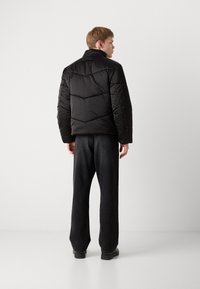 Black puffer jacket with a high collar, featuring a chevron pattern and a logo on the back. Worn over loose-fitting dark jeans.