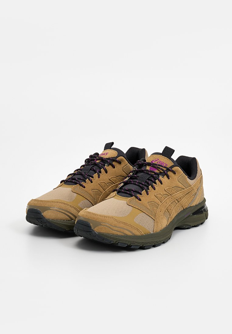 Une paire de baskets de sport beiges et olives avec des lacets noirs et violets, des semelles texturées et des cols rembourrés noirs sur fond blanc.