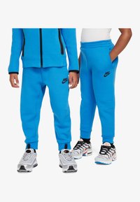 Sininen Nike huppari ja jogginghousut mustilla yksityiskohdilla, jossa on vetoketjukiinnitys ja logo. Yhdistetty harmaisiin ja mustiin lenkkareihin.