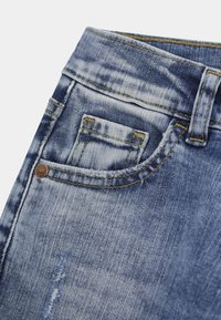 Pantalones vaqueros de mezclilla azul claro con parches desteñidos, un pequeño bolsillo delantero y costuras amarillas. Incluyen un detalle de logotipo metálico cerca del bolsillo.