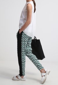 Donna che indossa una canottiera bianca senza maniche, pantaloni slim a fantasia con strisce laterali nere, sandali bianchi spessi e tiene una grande borsa nera.