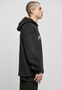 Schwarzer Kapuzenpullover mit lockerer Passform, weichem Material, Fronttasche und weißen Grafiken auf der Brust.